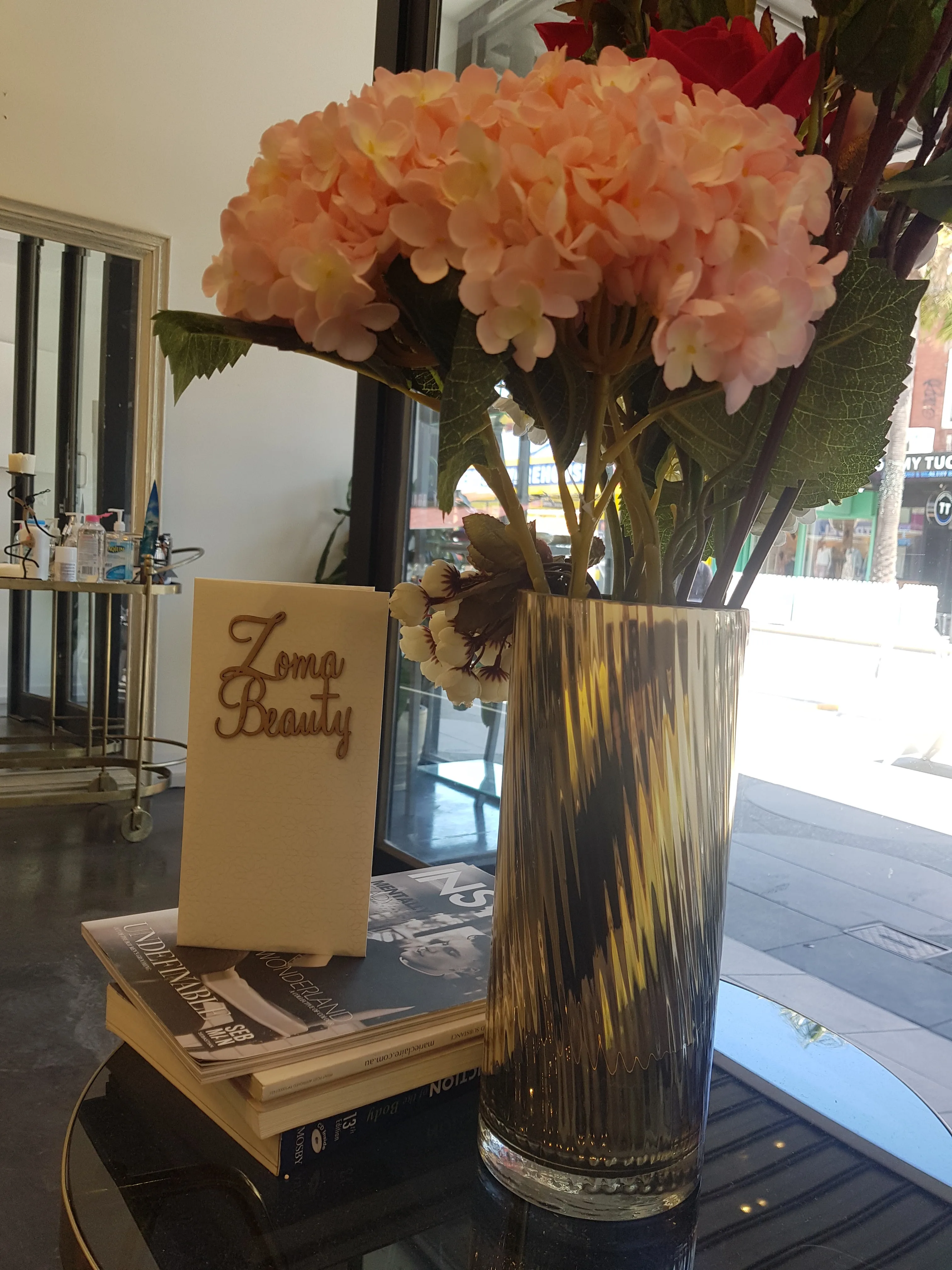 Window-side detail — fresh flowers beside the Zoma Beauty table card and magazines